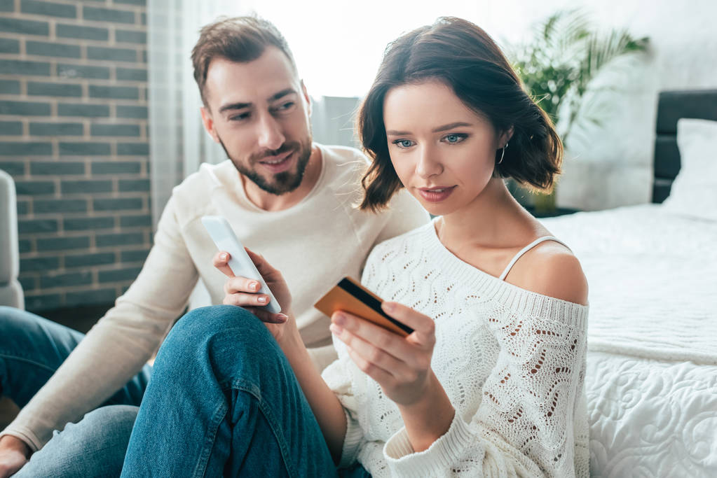 attractive woman holding smartphone and looking at credit card near cheerful man  - Photo, Image