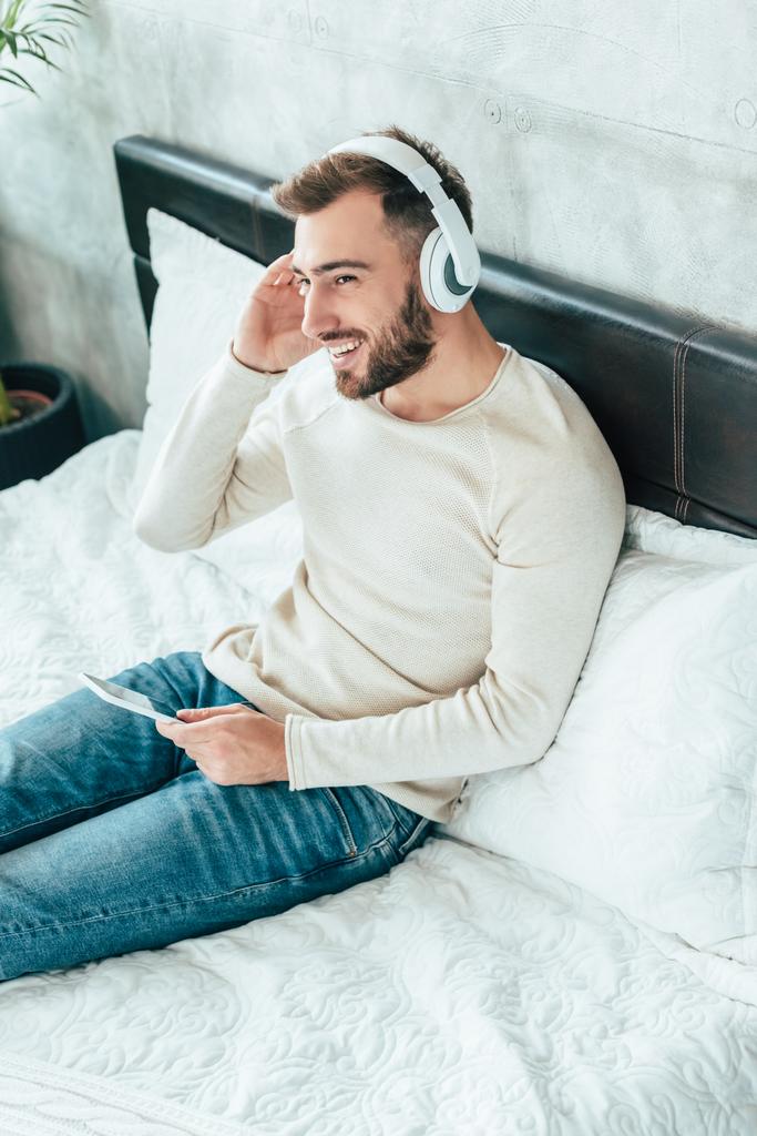 stropní pohled na šťastného vousatého muže, který používá smartphone při poslechu hudby ve sluchátkách  - Fotografie, Obrázek
