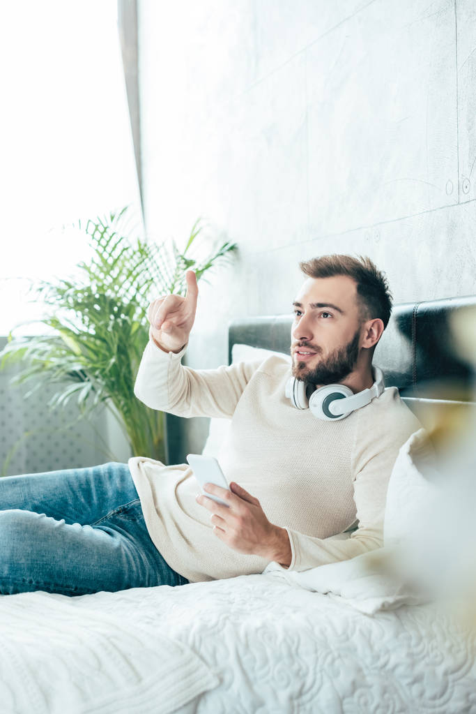 επιλεκτική εστίαση του όμορφου άντρα με γένια στα ακουστικά κρατώντας smartphone και δείχνοντας με το δάχτυλο στο υπνοδωμάτιο  - Φωτογραφία, εικόνα