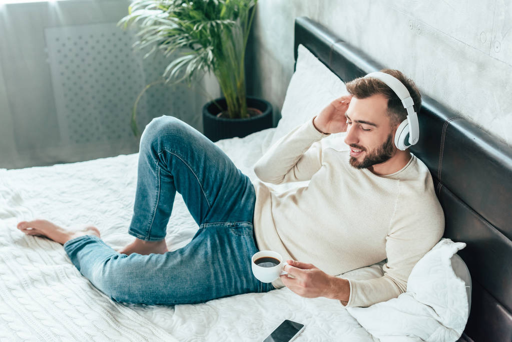 Widok z góry szczęśliwy człowiek słuchania muzyki w słuchawkach, trzymając filiżankę kawy w łóżku  - Zdjęcie, obraz