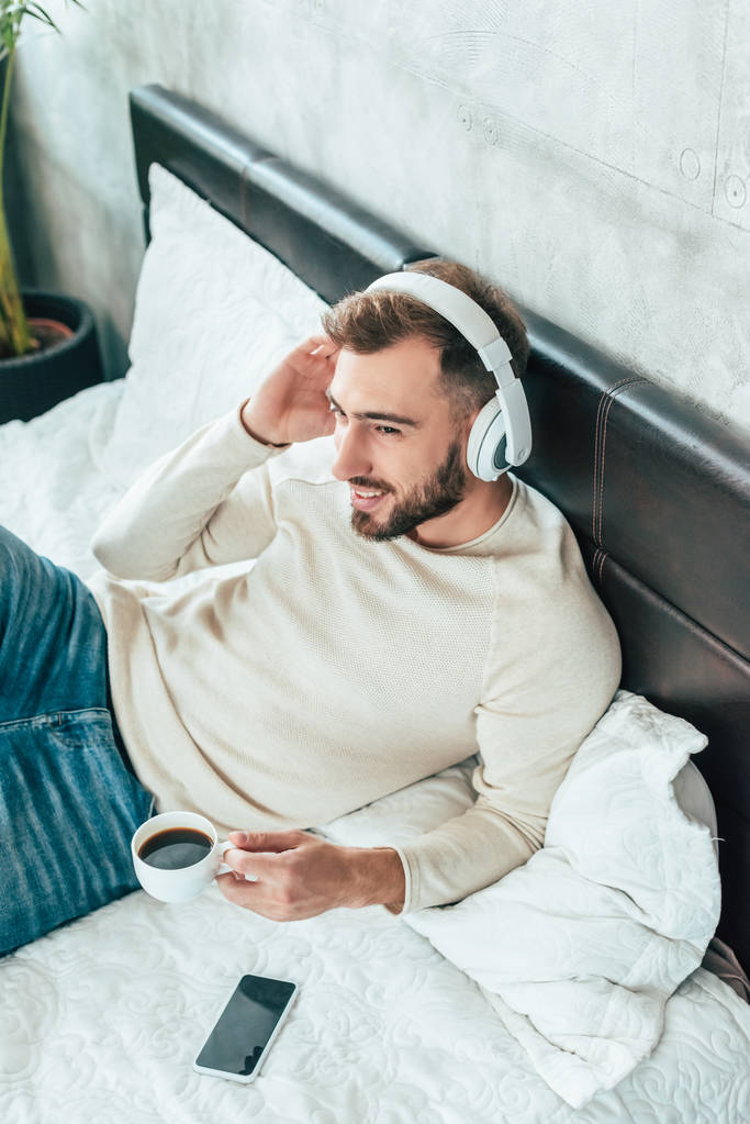ベッドでコーヒーを飲みながらヘッドフォンで音楽を聴く陽気な男の頭上の眺め  - 写真・画像