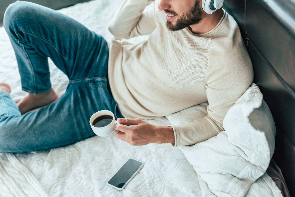 ベッドでコーヒーを飲みながらヘッドフォンで音楽を聴く陽気な男のトリミングされた景色  - 写真・画像