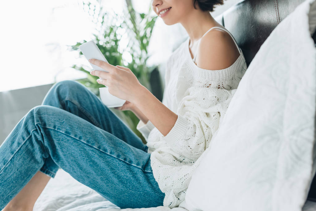 oříznutý pohled na šťastného ženu držící pohár a používání smartphone v ložnici  - Fotografie, Obrázek