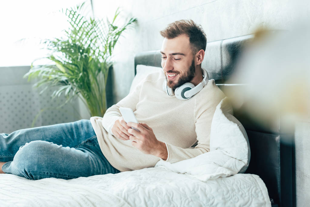 selektivní zaměření šťastného vousatého muže ve sluchátkách pomocí smartphone ve sluchátkách  - Fotografie, Obrázek