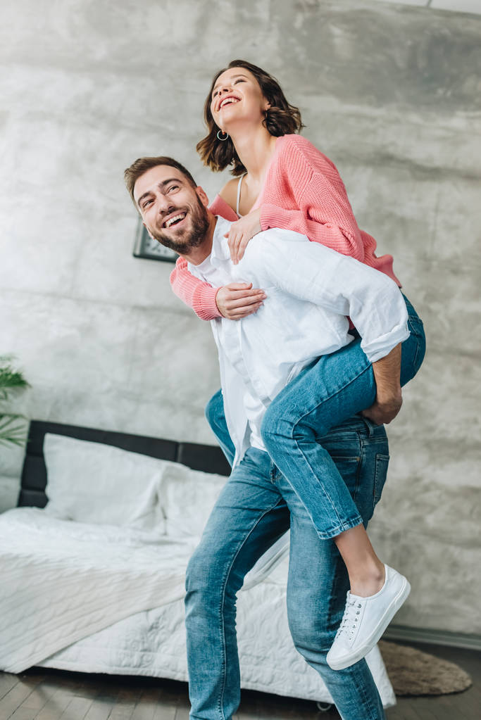 alacsony látószögű nézet boldog szakállas férfi piggybacking barna nő a hálószobában  - Fotó, kép
