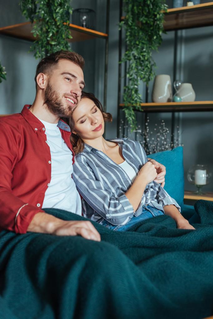 glücklicher Mann umarmt attraktive Frau mit geschlossenen Augen, während er zu Hause auf dem Sofa sitzt  - Foto, Bild