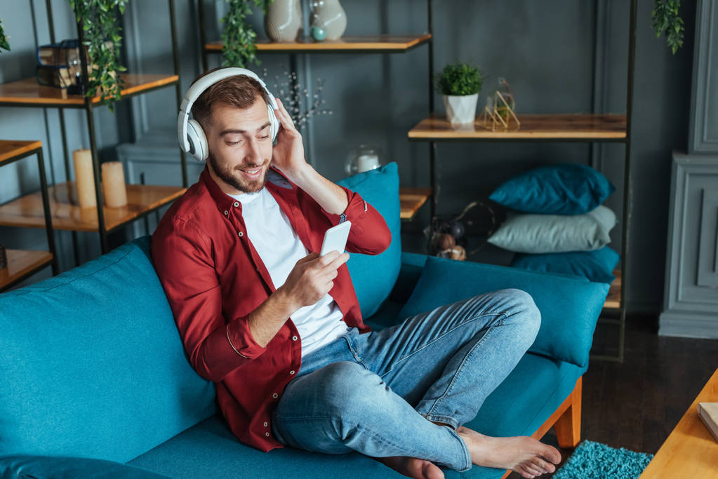 felice uomo barbuto ascoltare musica in cuffia e utilizzando smartphone in soggiorno
  - Foto, immagini