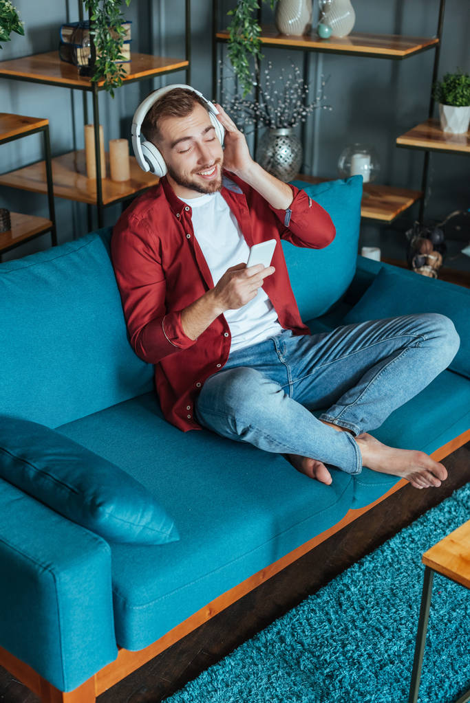 vista aérea del hombre barbudo feliz escuchando música en los auriculares y utilizando el teléfono inteligente en la sala de estar
  - Foto, Imagen