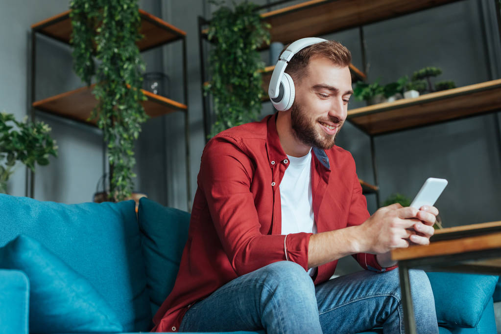 vista a basso angolo di felice uomo barbuto ascoltare musica in cuffia e utilizzando smartphone in soggiorno
  - Foto, immagini