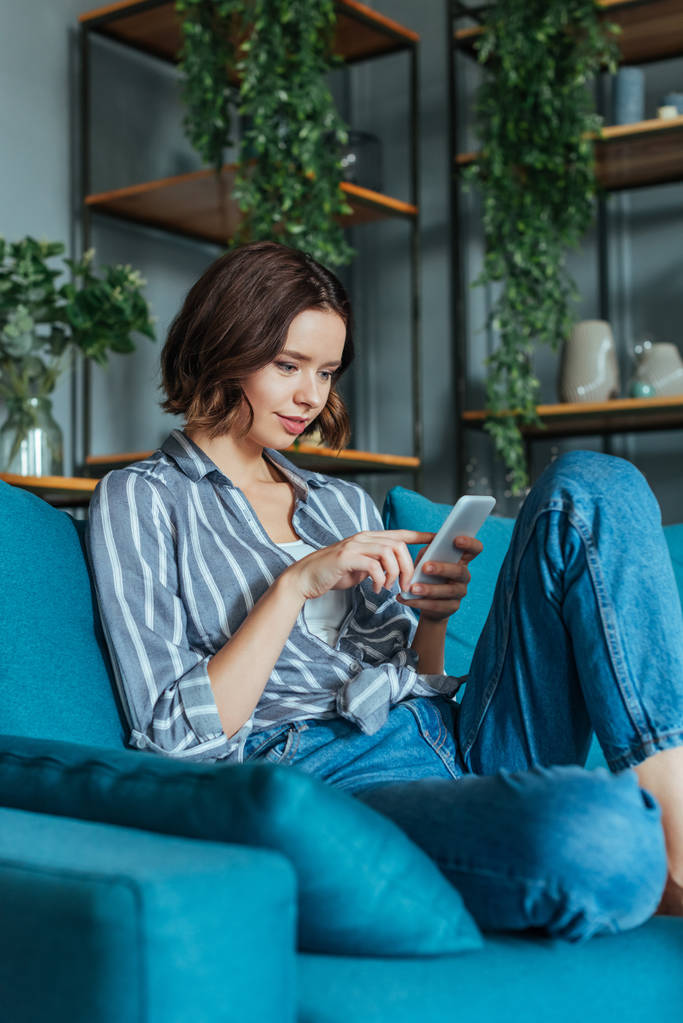 pohled s nízkým výhledem na atraktivní brunetovou ženu používající smartphone v obývacím pokoji  - Fotografie, Obrázek