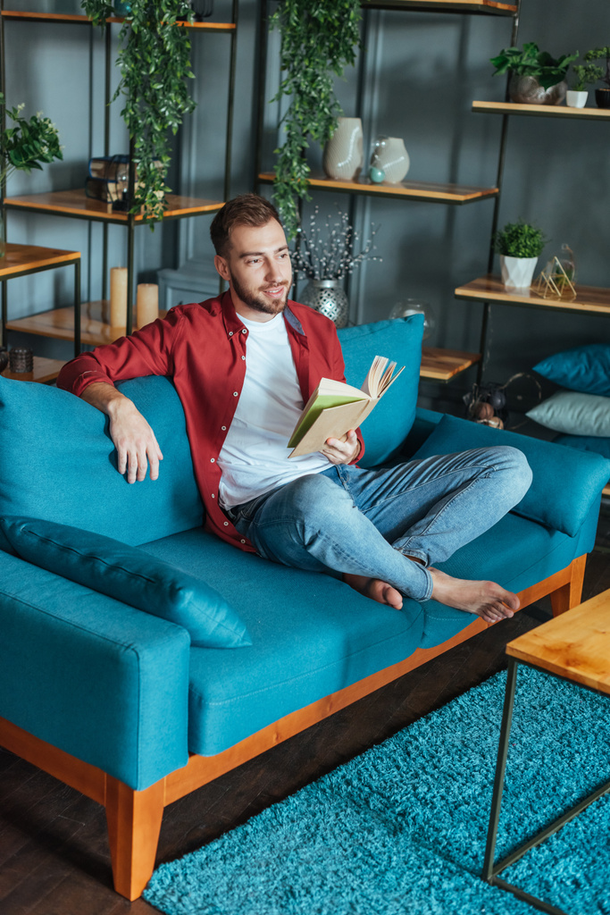 jóképű ember ül a kanapén és a gazdaság könyv a nappaliban  - Fotó, kép