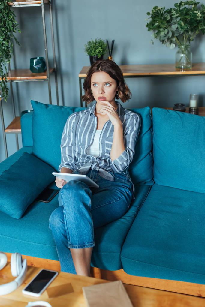 pensive brunette woman holding digital tablet while sitting in living room  - Photo, Image