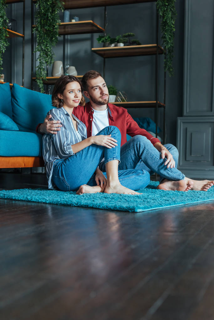 pohled nízkého úhlu šťastného muže, který se při sezení na koberci v obývacím pokoji objímat s atraktivní ženou  - Fotografie, Obrázek