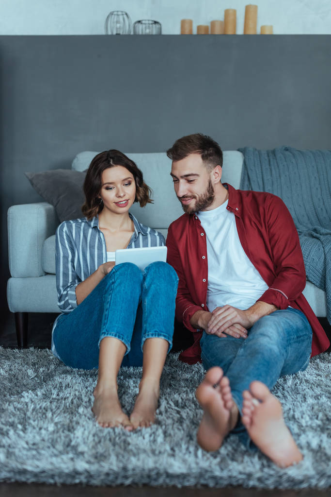 glücklicher Mann und Frau auf Teppich sitzend und auf digitales Tablet blickend  - Foto, Bild