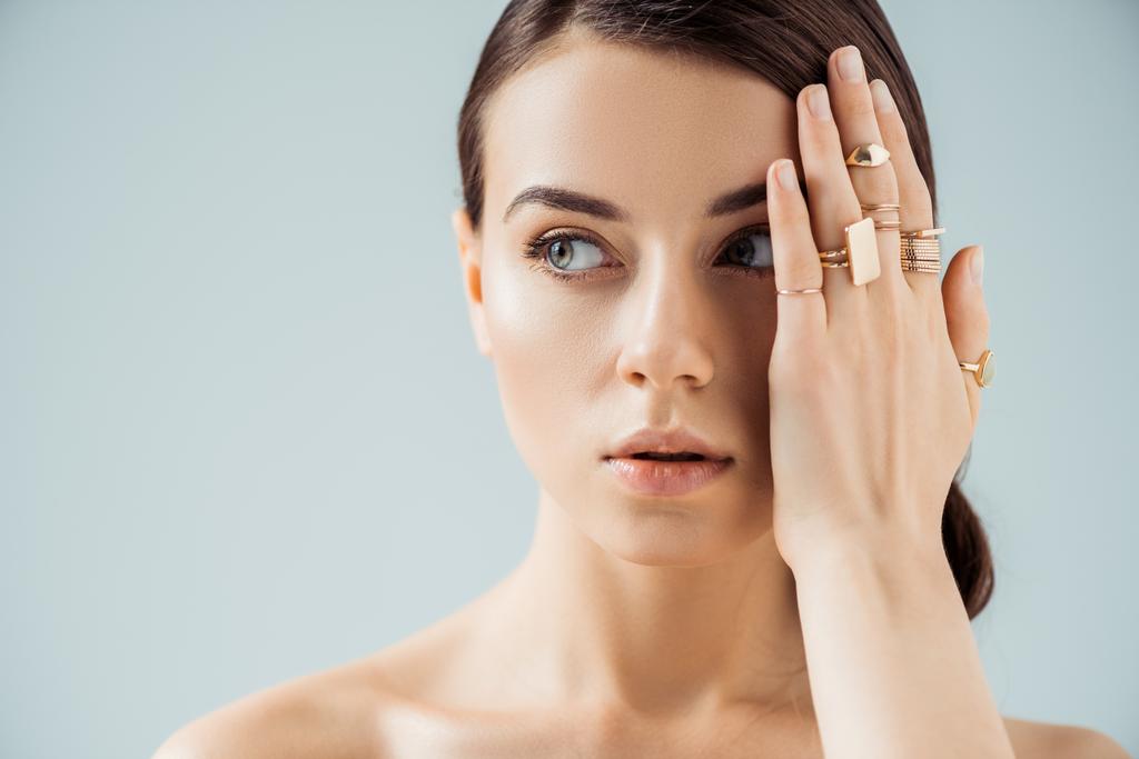 young naked woman with shiny makeup and golden rings hiding face behind hand and looking away isolated on grey - Photo, Image
