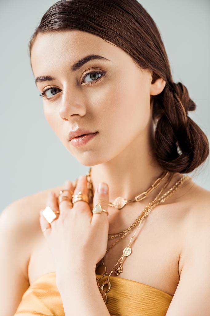 młoda atrakcyjna kobieta z błyszczącym makijażem w złotych naszyjnikach i pierścieniach patrząc na aparat odizolowany na szarości - Zdjęcie, obraz