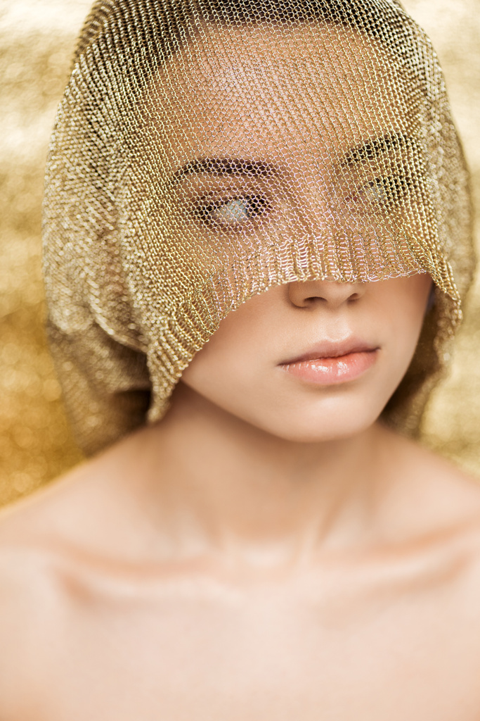 νεαρή γυμνή γυναίκα με λαμπερά χείλη και χρυσό πανί στο πρόσωπο κοιτάζοντας μακριά σε χρυσό υφή φόντο - Φωτογραφία, εικόνα