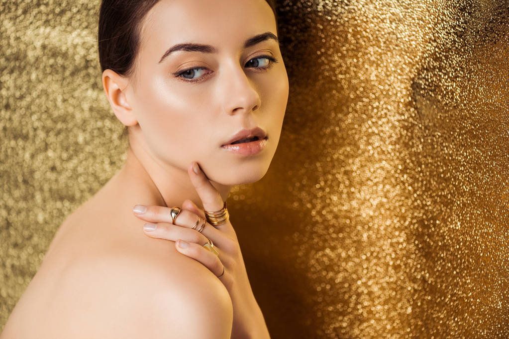 mujer joven desnuda con maquillaje brillante y anillos de oro mirando hacia otro lado sobre fondo de textura dorada
 - Foto, imagen