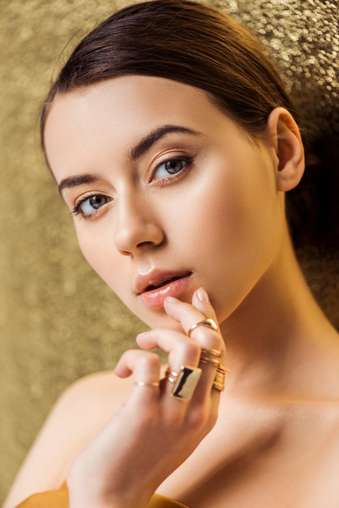 atrakcyjna młoda kobieta z błyszczącym makijażu i złote pierścienie dotykając usta na złotym tle teksturowane - Zdjęcie, obraz