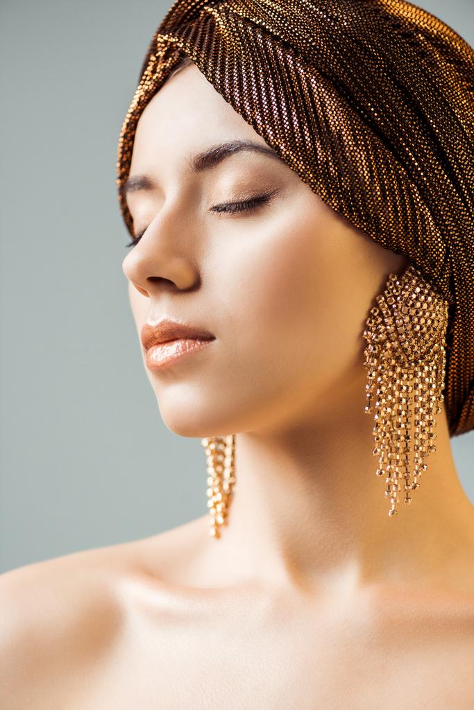 mladá nahá žena se zavřenýma očima, lesklý make-up, zlaté prsteny v turbanu izolované na šedi - Fotografie, Obrázek