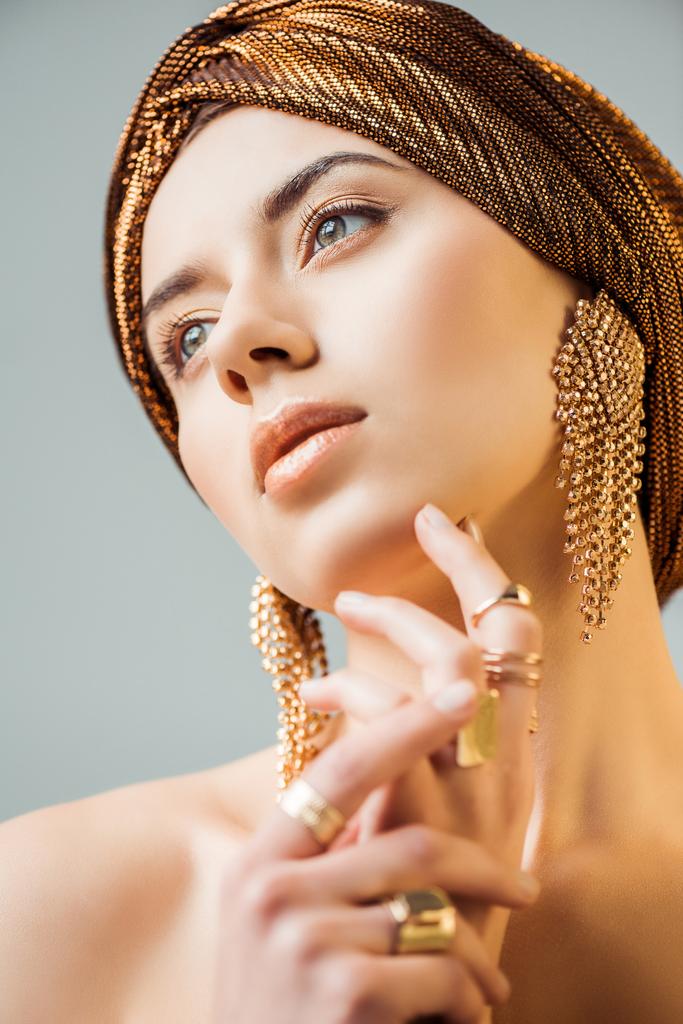 junge nackte Frau mit glänzendem Make-up, goldenen Ringen und Ohrringen in Turban isoliert auf grau - Foto, Bild