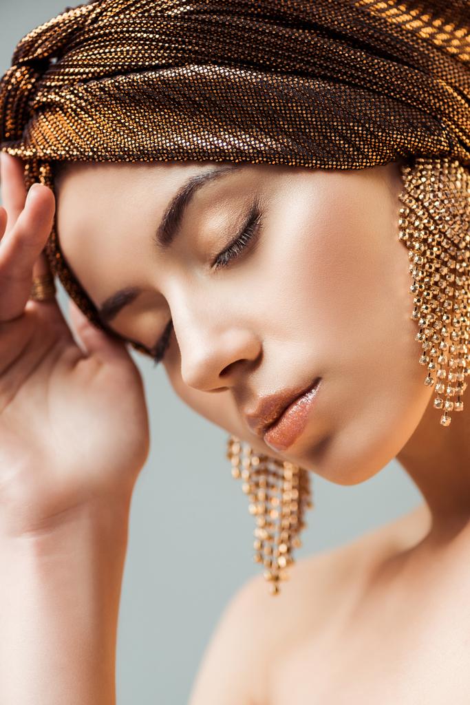 молодая женщина с закрытыми глазами, блестящий макияж и золотые серьги в тюрбане изолированы на сером
 - Фото, изображение