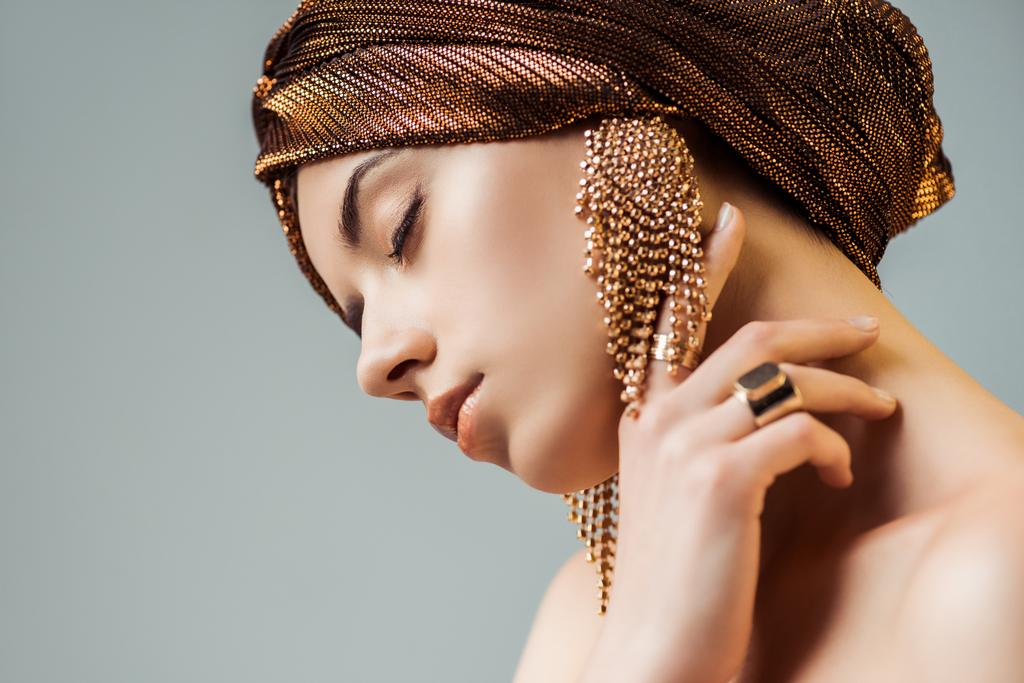 young nude woman with closed eyes, shiny makeup, golden rings and earrings in turban touching neck isolated on grey - 写真・画像