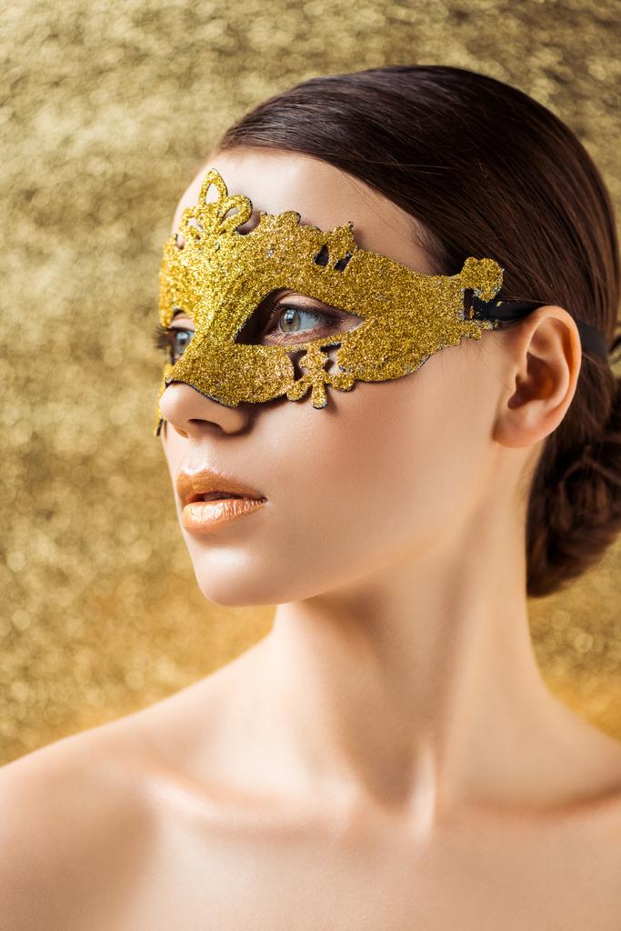 νεαρή γυμνή γυναίκα σε χρυσή μάσκα με λαμπερό μακιγιάζ κοιτάζοντας μακριά σε υφή χρυσό φόντο - Φωτογραφία, εικόνα