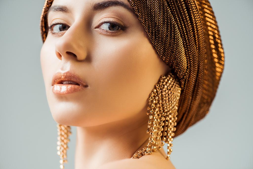 junge schöne Frau mit glänzendem Make-up, Turban und goldenen Ohrringen, die in die Kamera schaut, isoliert auf grau - Foto, Bild