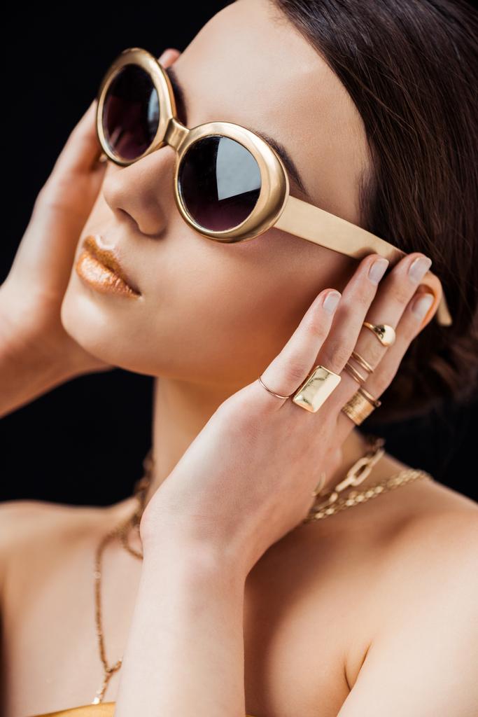 mladá žena ve slunečních brýlích, zlatých prstenech a náhrdelnících dotýkajících se uší izolovaných na černé - Fotografie, Obrázek