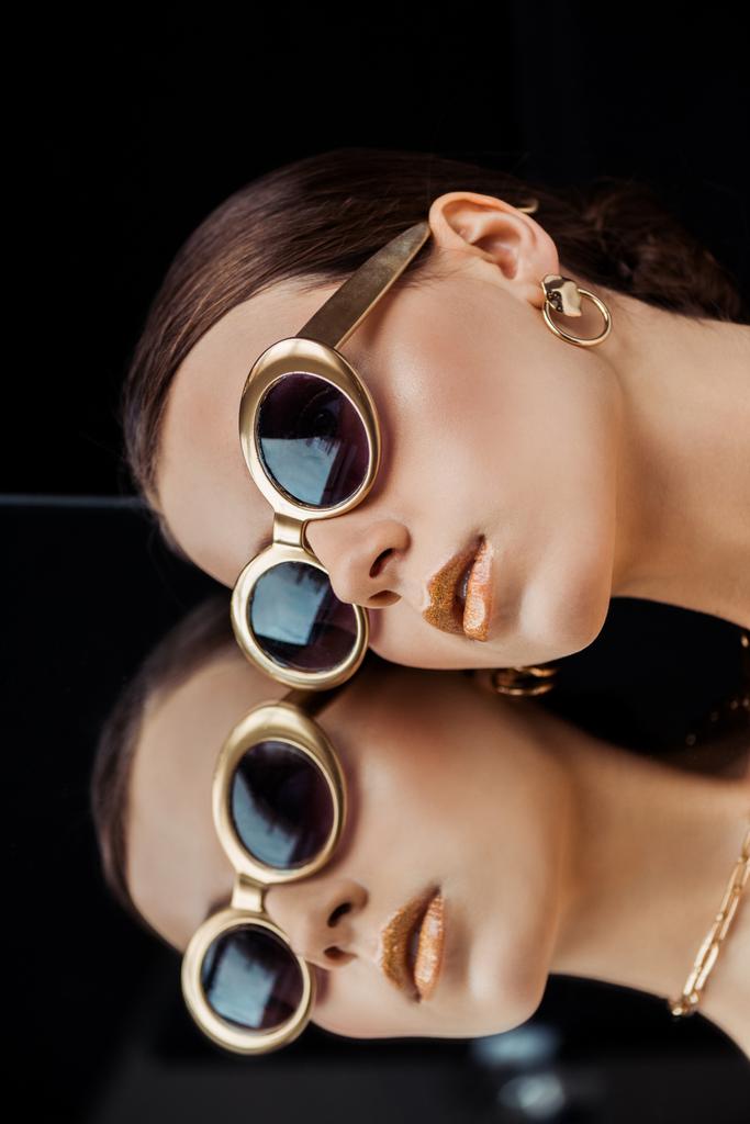 jovem mulher em óculos de sol, jóias de ouro deitado no espelho isolado em preto
 - Foto, Imagem