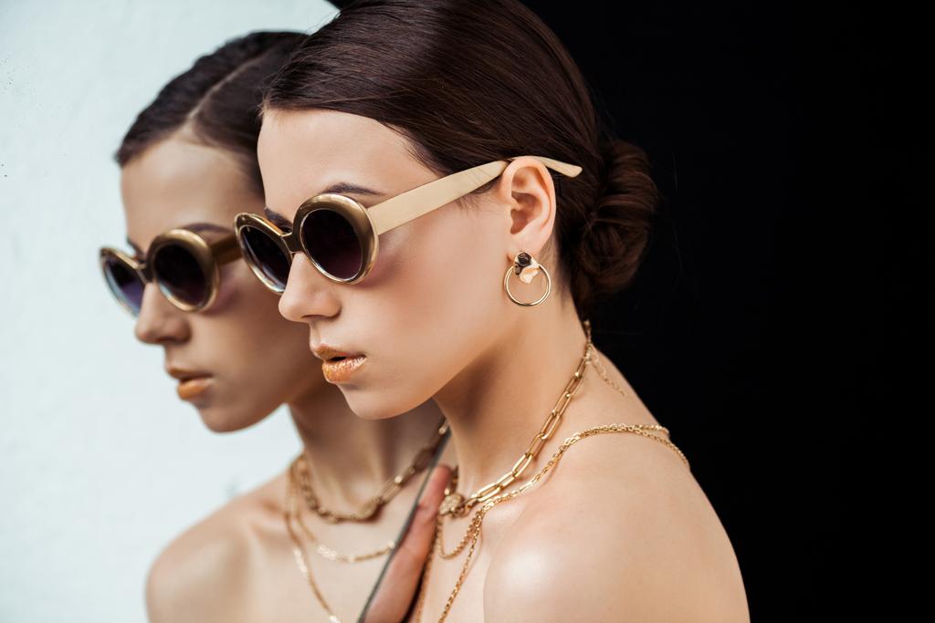 jonge naakte vrouw in zonnebril, gouden accessoires houden spiegel geïsoleerd op zwart - Foto, afbeelding