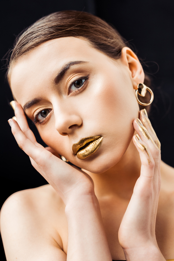 νεαρή γυμνή γυναίκα με χρυσό μακιγιάζ και χρυσό χρώμα στα δάχτυλα αγγίζοντας το πρόσωπο απομονώνονται σε μαύρο - Φωτογραφία, εικόνα
