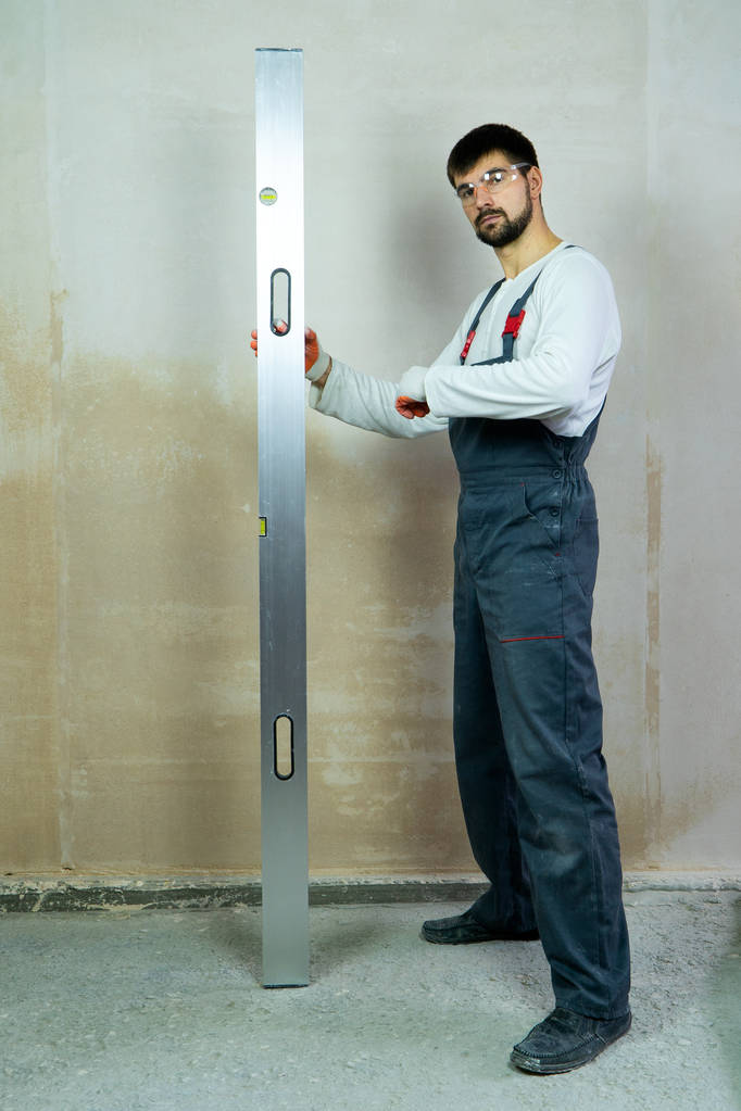 young bearded builder stands near empty wall with construction ruler - Photo, Image