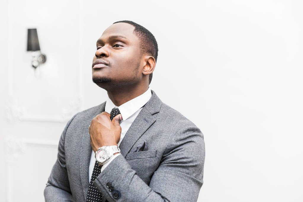 Nuori afroamerikkalainen liikemies harmaassa puvussa sitomassa solmiota valkoisella taustalla
. - Valokuva, kuva