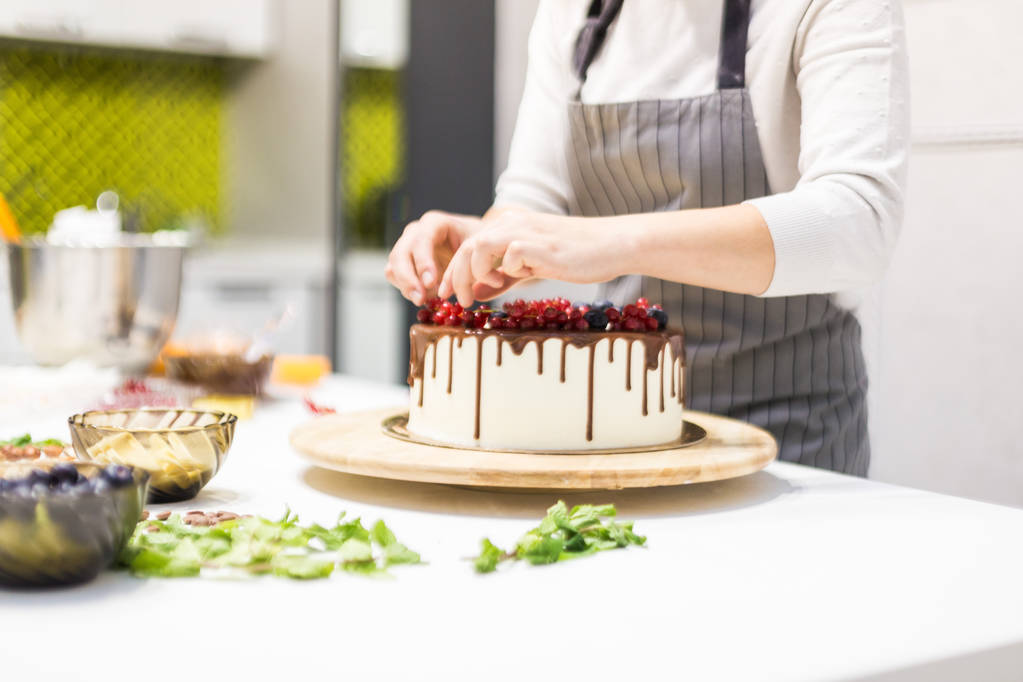 Кондитер украшает ягодами бисквитный торт с белым кремом и шоколадом. Торт стоит на деревянном столике на белом столе. Концепция домашней выпечки, приготовления тортов
. - Фото, изображение