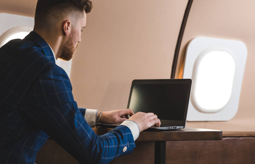 Atrakcyjny i udany biznesmen pracujący na laptopie siedząc na krześle swojego prywatnego odrzutowca. - Zdjęcie, obraz