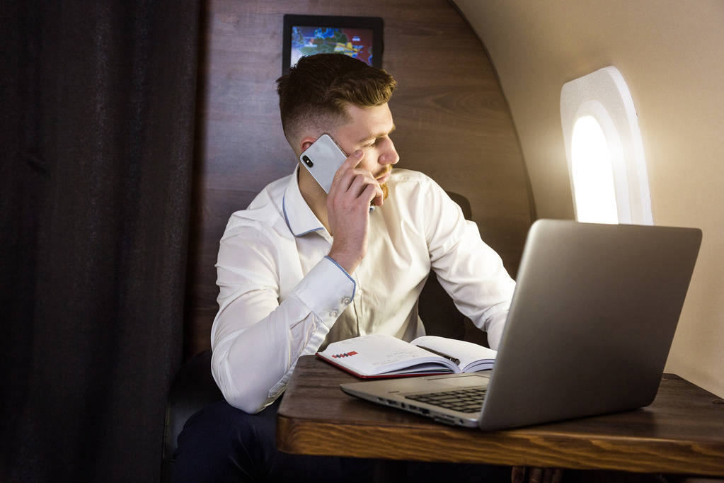 Joven empresario atractivo y exitoso hablando por teléfono y trabajando mientras está sentado en la silla de su avión privado de negocios
 - Foto, Imagen