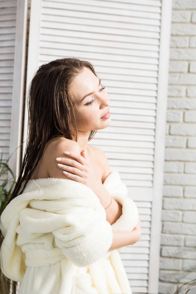 Ελκυστικό νεαρό κορίτσι με λευκό μπουρνούζι απλώνει κρέμα στο δέρμα ενώ κάθεται στο εσωτερικό του μπάνιου και κάνει πρωινές επεμβάσεις. Φροντίδα δέρματος και σώματος - Φωτογραφία, εικόνα