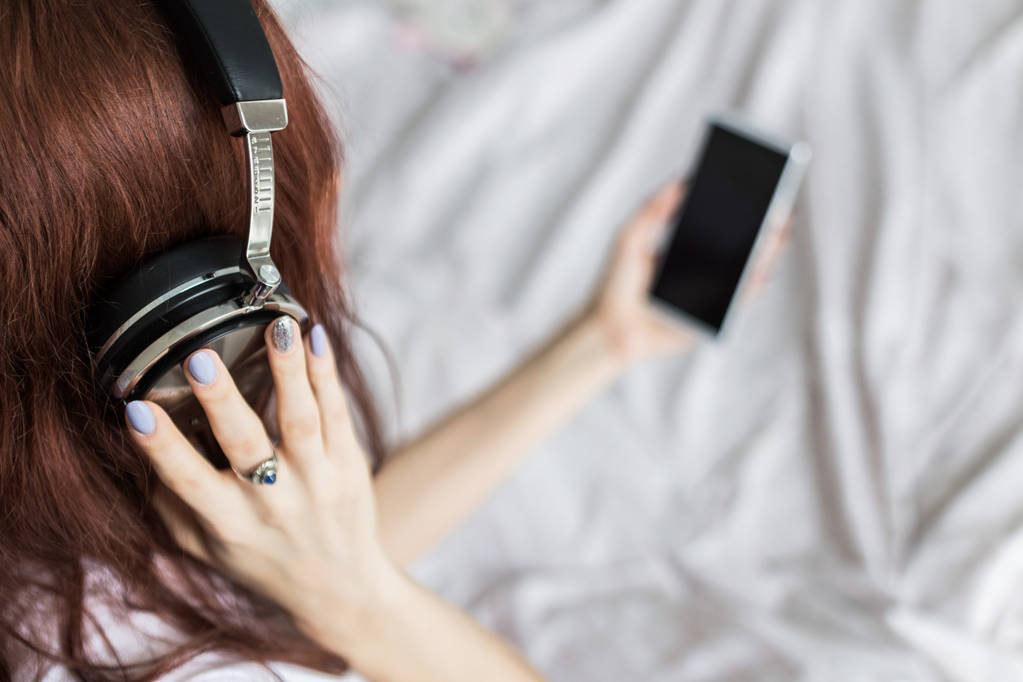 Atraktivní mladá brunetka žena sedí ráno ve své posteli se sluchátky a smartphone a poslouchá hudbu v dobré náladě - Fotografie, Obrázek