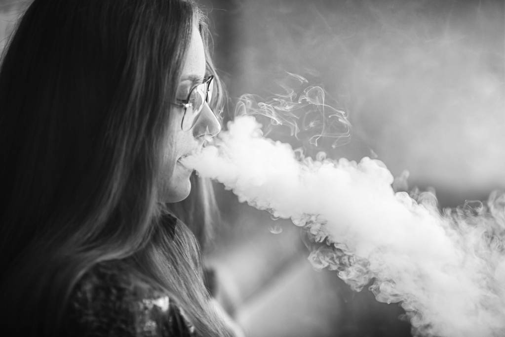 Подросток с проблемной кожей. Портрет молодой милой девушки в солнечных очках, курящей электронную сигарету в баре. Плохая привычка, вредная для здоровья. Падающая активность. Черное над белым. Закрыть
. - Фото, изображение