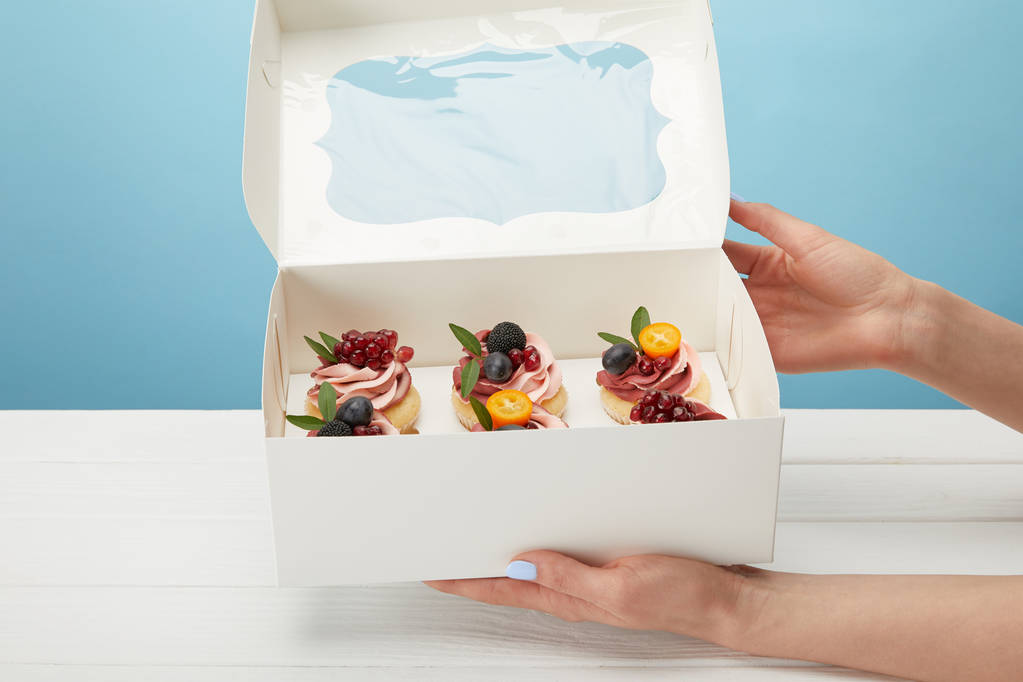 青で隔離されたカップケーキを持つ箱を持つ女性の部分的なビュー - 写真・画像