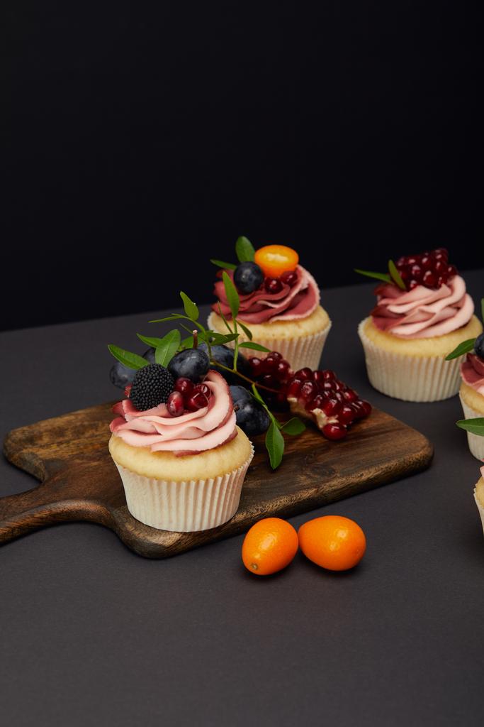 sweet cupcakes with fruits and berries and cutting board on grey surface isolated on black - Photo, Image