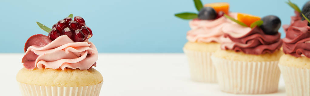panoramic shot of cupcakes with cream and garnet on white surface isolated on blue - Photo, Image
