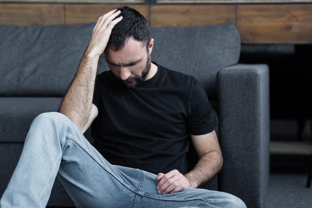 masentunut parrakas mies istuu lattialla lähellä harmaa sohva ja pitää kädestä pään
 - Valokuva, kuva