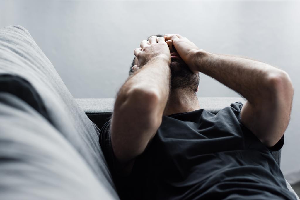 Depressiver Mann liegt auf Sofa und leidet, während er die Hände vor dem Gesicht hält - Foto, Bild