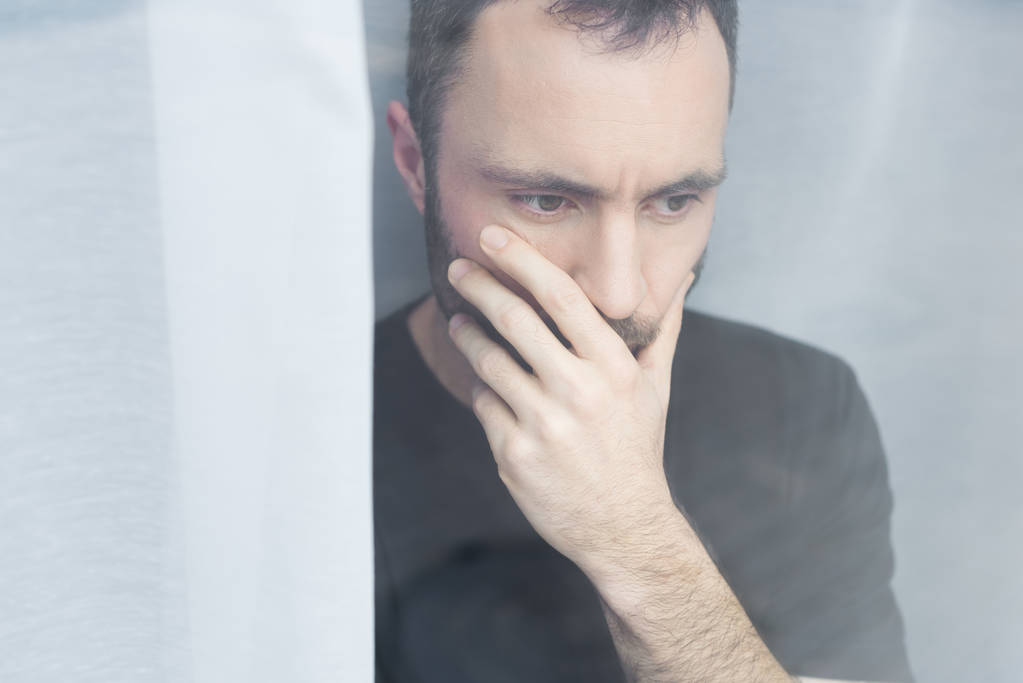 handsome man in black t-shirt standing by window and holding hand on mouth - Photo, Image