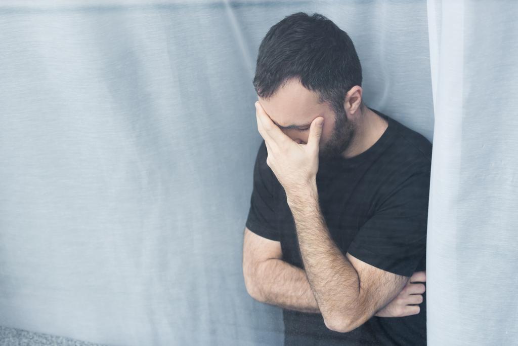 lonely man in black t-shirt standing by window at home and holding hand on face - Photo, Image
