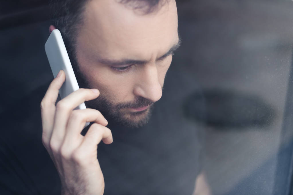 upset handsome man standing by window with smartphone and looking away - Photo, Image