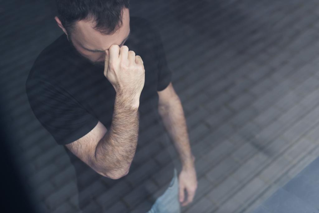 hoge hoek weergave van depressieve man in zwart t-shirt staand door venster en holding hand in de buurt van gezicht - Foto, afbeelding